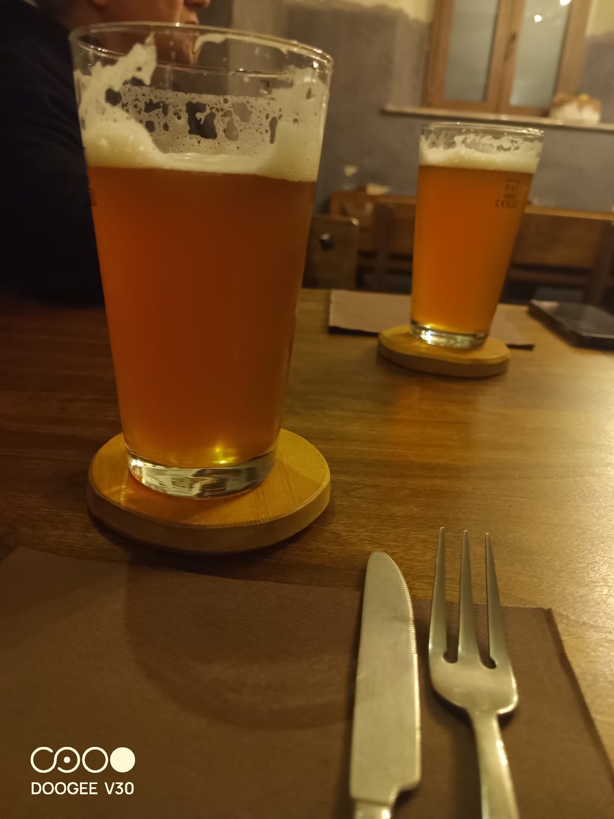 Due birre artigianali ambrate al tavolo, su sottobicchieri di legno, con posate su una tovaglietta.