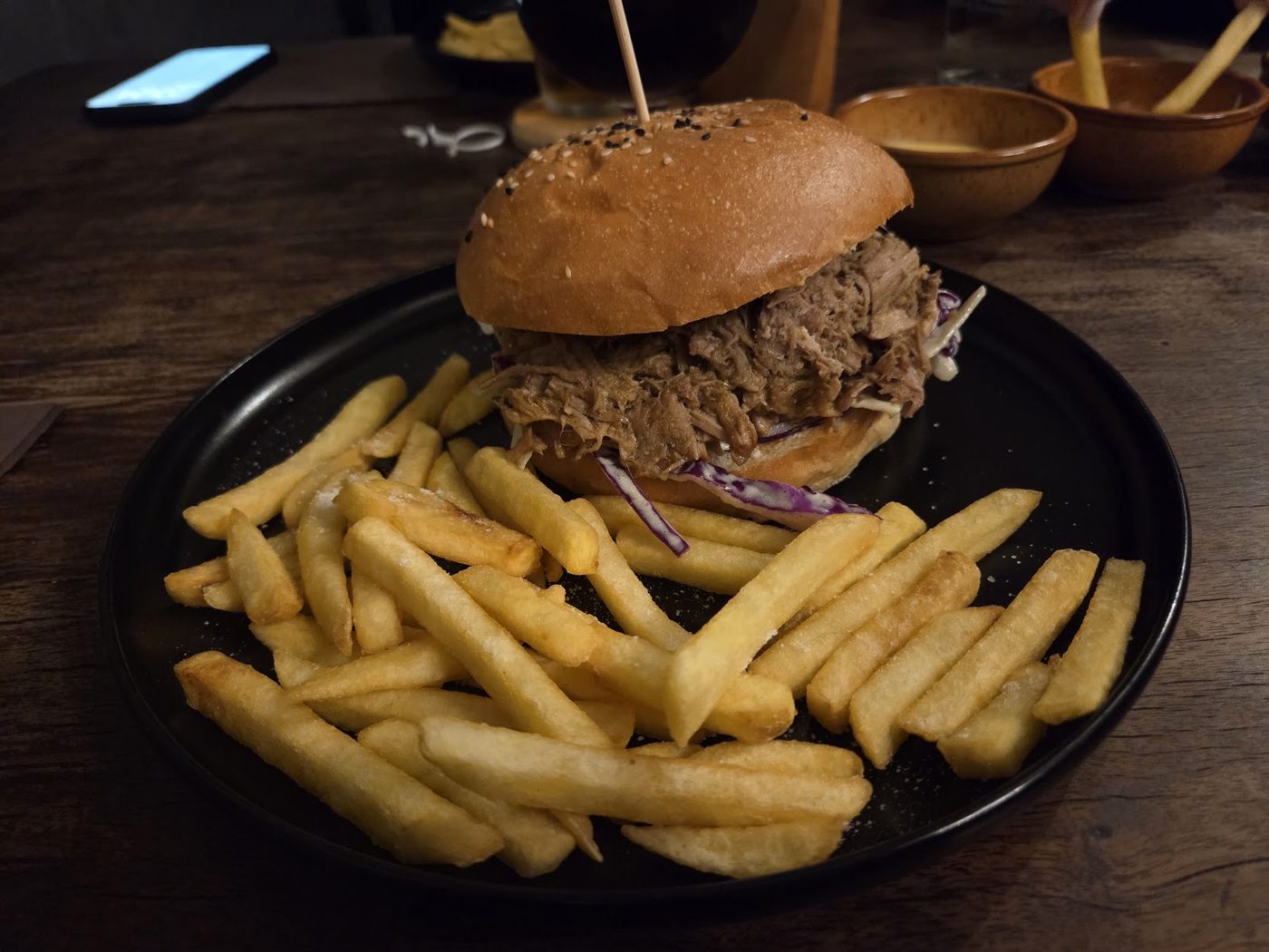Hamburger di maiale sfilacciato con cavolo rosso in un panino ai semi, con patatine e salse in ciotole di terracotta.