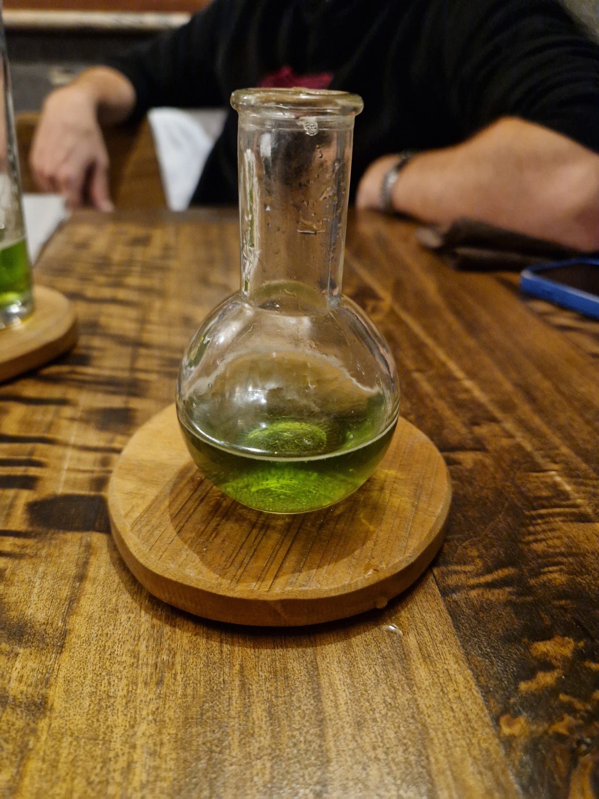 Un liquore verde servito in un&rsquo;ampolla da laboratorio su un sottobicchiere di legno.