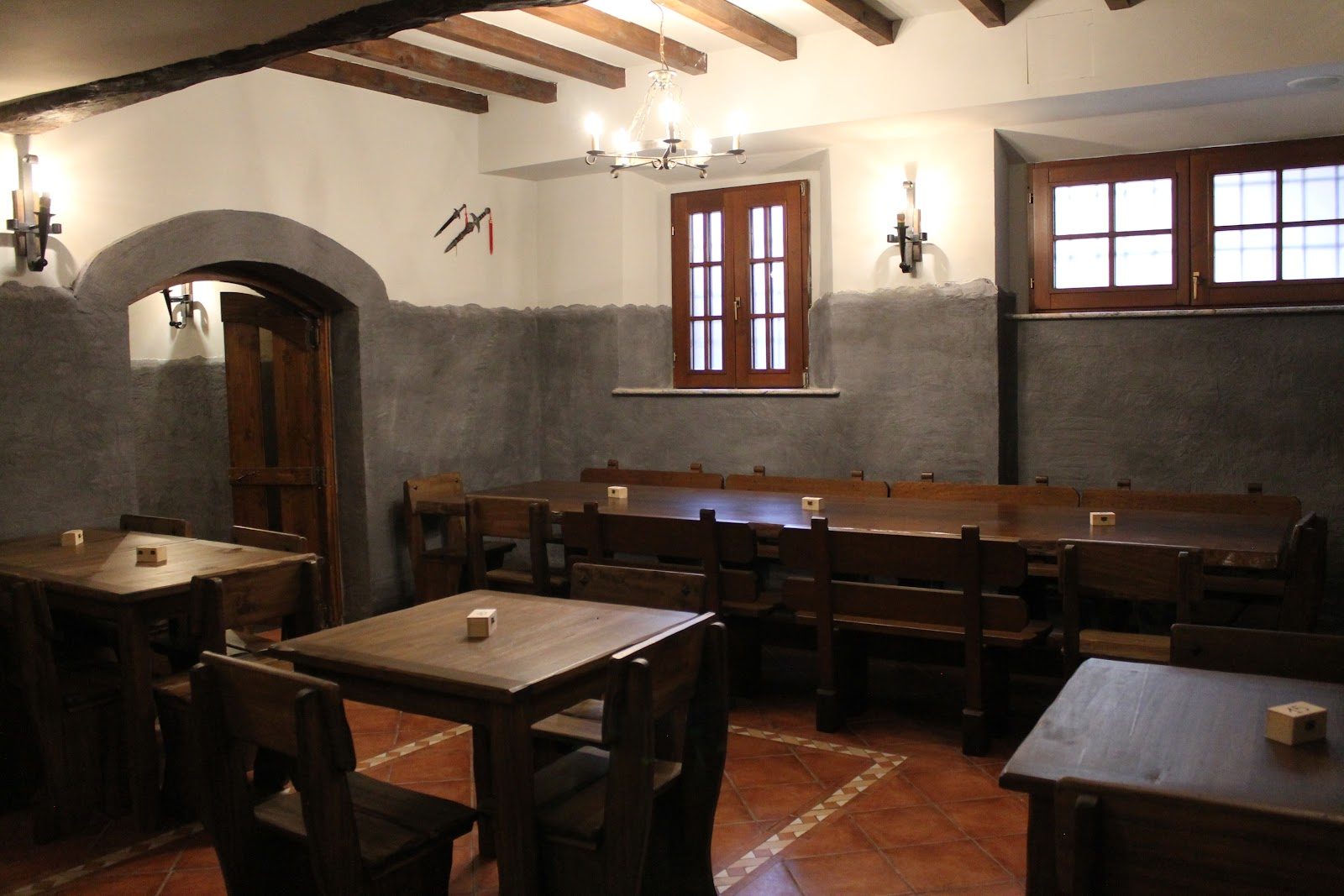 Una sala vuota della taverna: zoccolatura in intonaco grigio, arco in pietra, tavolata lunga con panche, pugnali appesi alla parete.