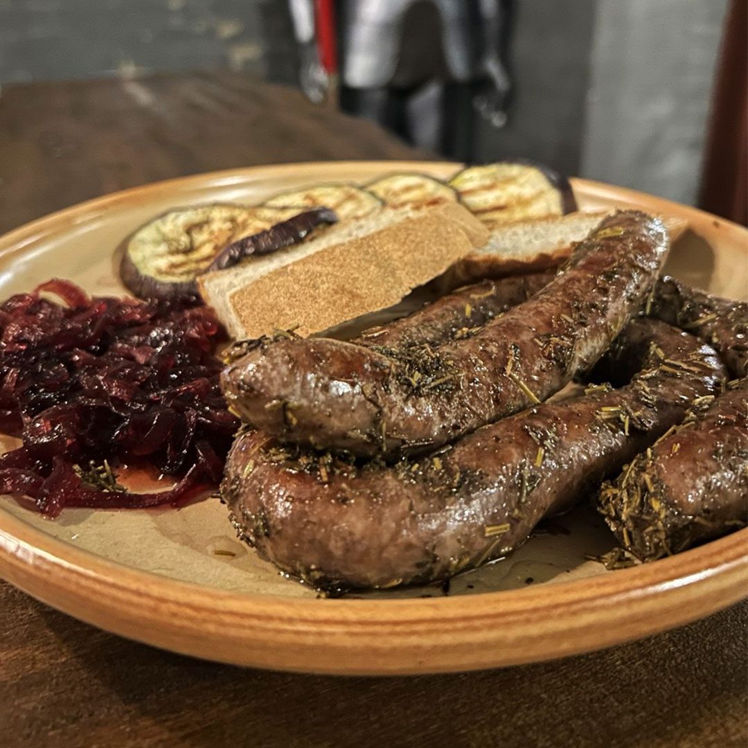 Salsicce al rosmarino con cipolle al vino rosso, melanzana grigliata e pane, servite su un piatto di terracotta.