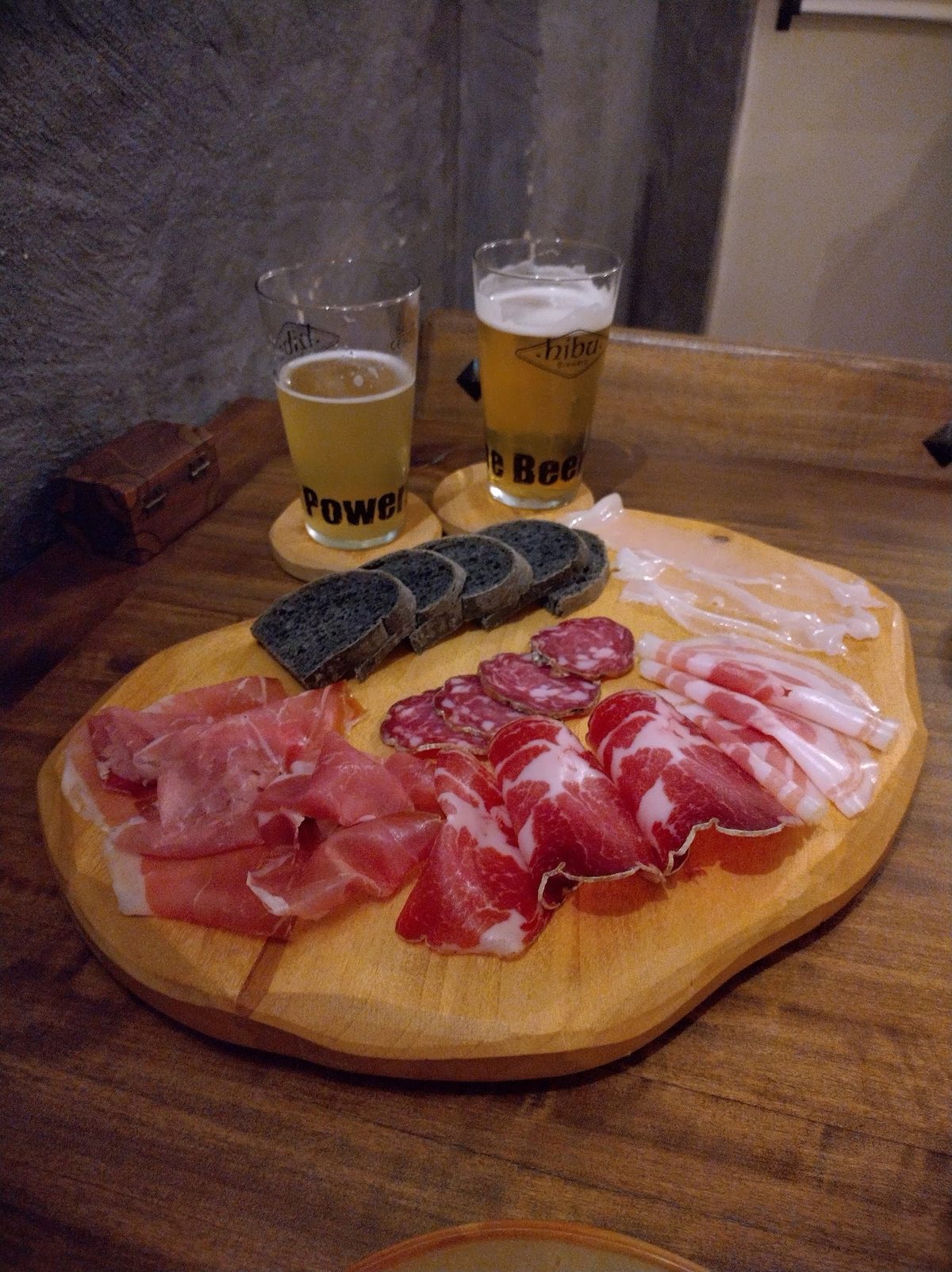 Tagliere di salumi affettati con pane al carbone e due birre artigianali su sottobicchieri di legno.